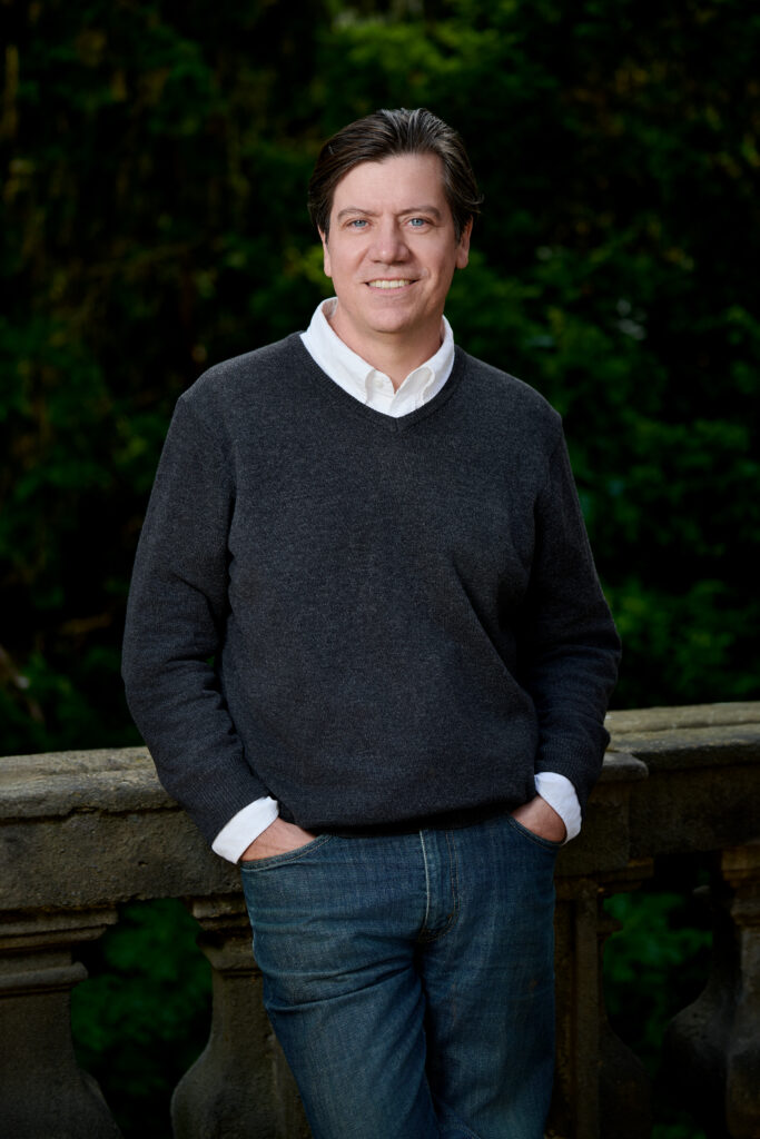 A photograph of Evan Rail wearing jeans and a gray v-neck sweater over a white shirt. 